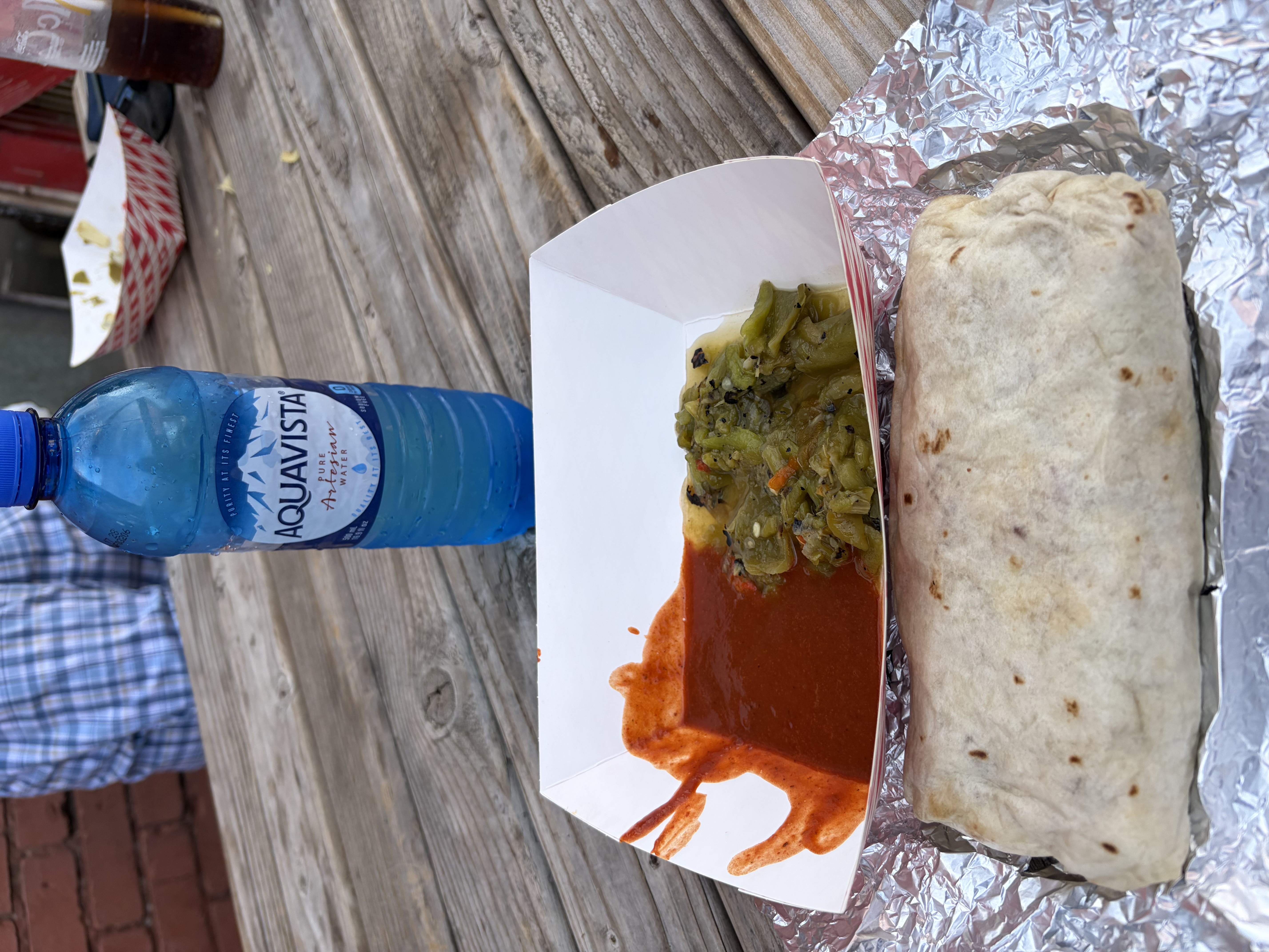 A foil-wrapped burrito on a picnic table beside a paper tray holding red chile sauce and chopped roasted green chile.